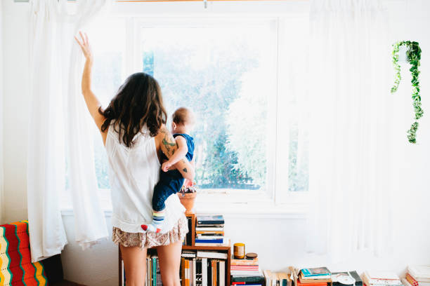 Jeune maman avec son bébé dans les bras qui ouvre la fenêtre pour aérer la chambre à cause des acariens